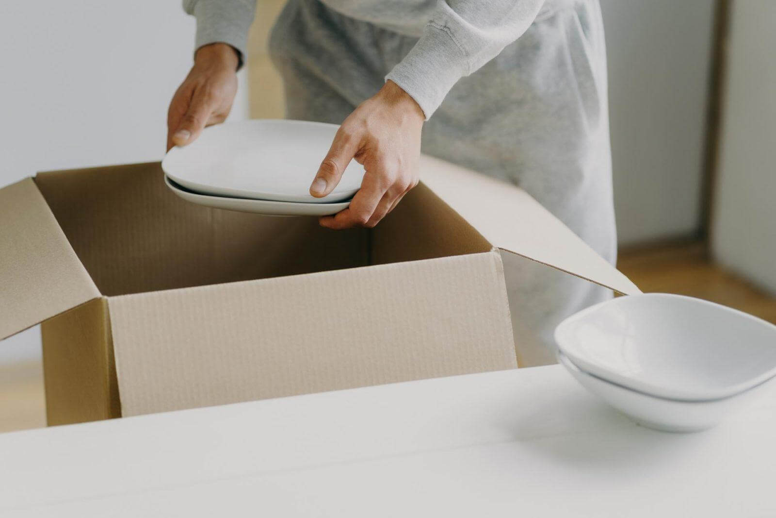 Busy man unpacks boxes with dishes, holds white plates, moves in new apartment, tidies up at kitchen, has moving day.